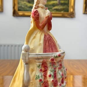 May include: A porcelain figurine of a woman in a yellow and red dress, standing on a balcony decorated with red roses. The figurine has blonde hair and is looking upwards. The balcony is white and has a small ball on top of the railing.