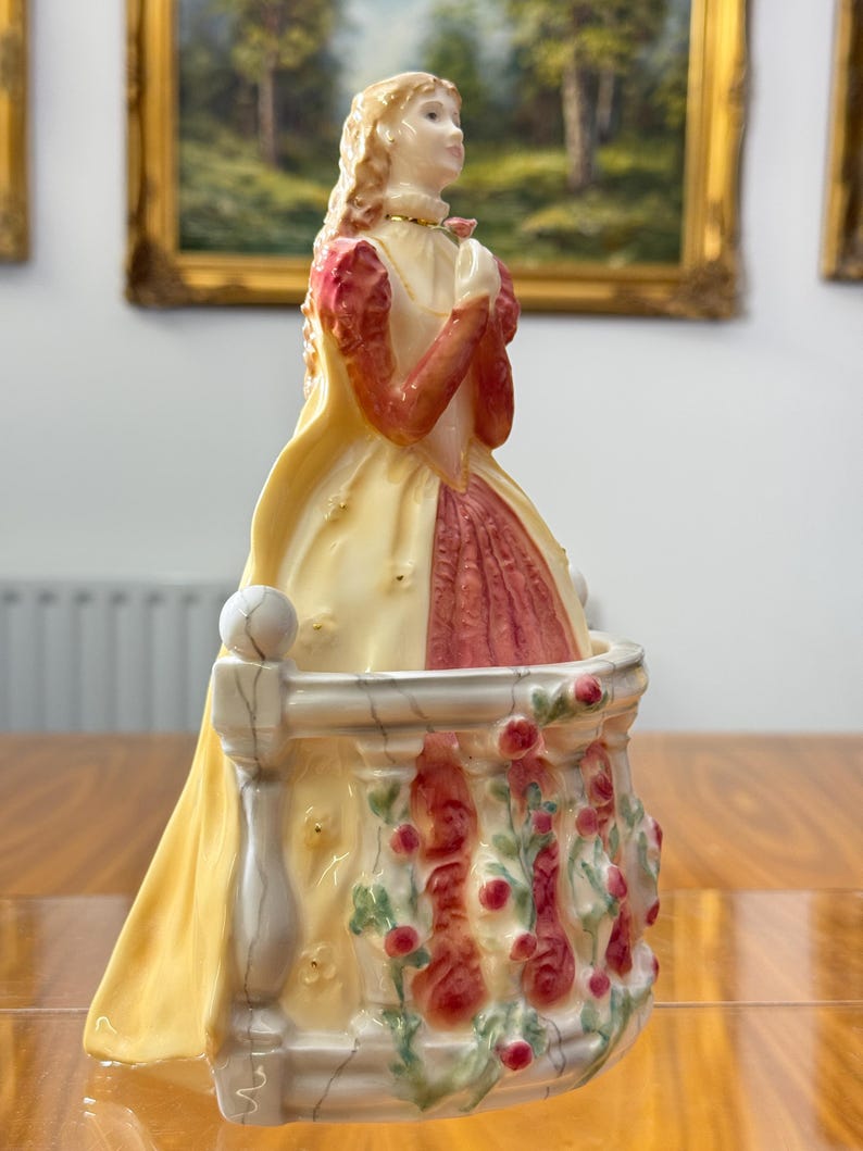 May include: A porcelain figurine of a woman in a yellow and red dress, standing on a balcony decorated with red roses. The figurine has blonde hair and is looking upwards. The balcony is white and has a small ball on top of the railing.