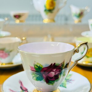 May include: A vintage teacup and saucer set with a floral rose design. The teacup features a gold handle and base, with a pink interior. The saucer and cup are decorated with a burgundy rose and green leaves. A teapot and other teacups are in the background.