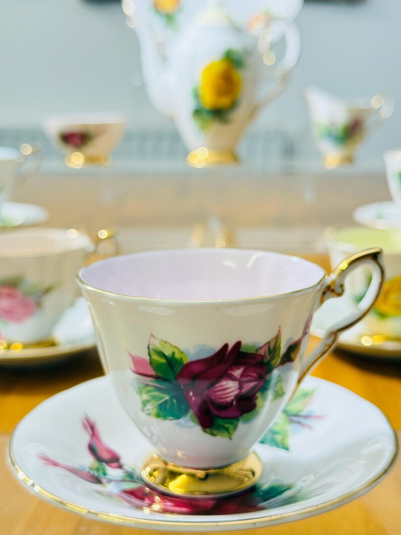 May include: A vintage teacup and saucer set with a floral rose design. The teacup features a gold handle and base, with a pink interior. The saucer and cup are decorated with a burgundy rose and green leaves. A teapot and other teacups are in the background.