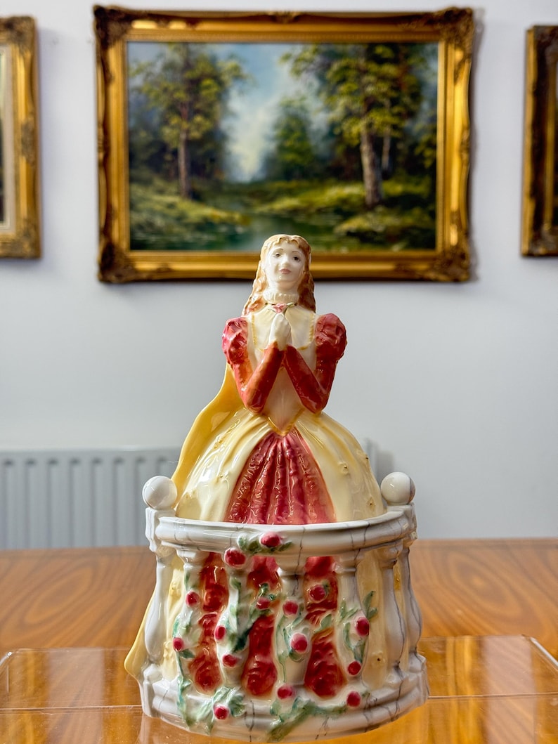 May include: A vintage porcelain figurine of a woman in a yellow and red dress, hands clasped in prayer. The figurine sits atop a decorative base with red rose accents. The background features framed landscape paintings.