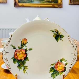 May include: A white porcelain serving plate with a scalloped edge and gold trim. The plate features hand-painted yellow and orange roses with green leaves. The plate is displayed on a gold-colored metal stand.