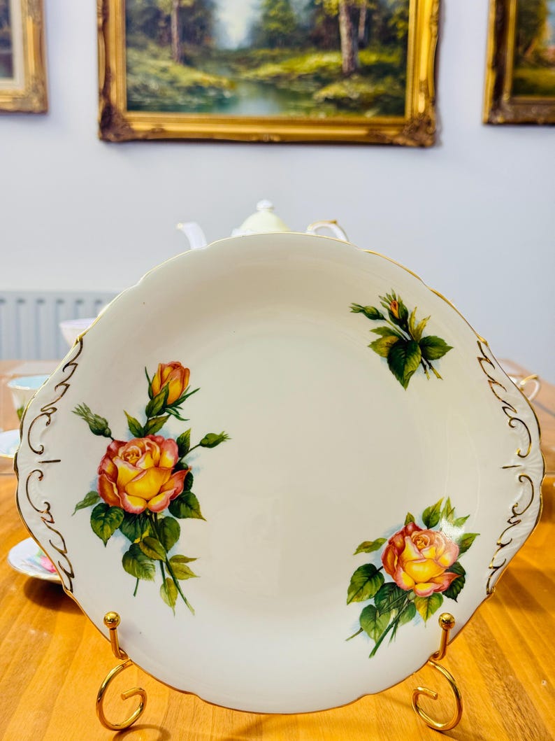 May include: A white porcelain serving plate with a scalloped edge and gold trim. The plate features hand-painted yellow and orange roses with green leaves. The plate is displayed on a gold-colored metal stand.
