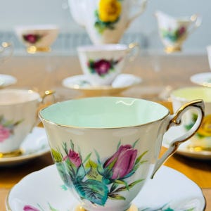 May include: A vintage tea set featuring a teacup with a light green interior, a saucer, and a gold-trimmed handle. The set is decorated with pink roses and green leaves. A teapot and other teacups are visible in the background.