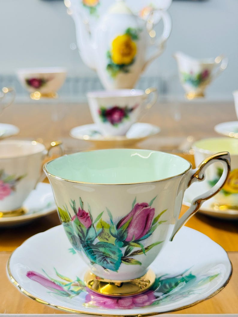 May include: A vintage tea set featuring a teacup with a light green interior, a saucer, and a gold-trimmed handle. The set is decorated with pink roses and green leaves. A teapot and other teacups are visible in the background.