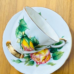 May include: White teacup and saucer set with a floral rose design in yellow and pink, accented with green leaves. The teacup and saucer have gold trim. The set is placed on a wooden surface.
