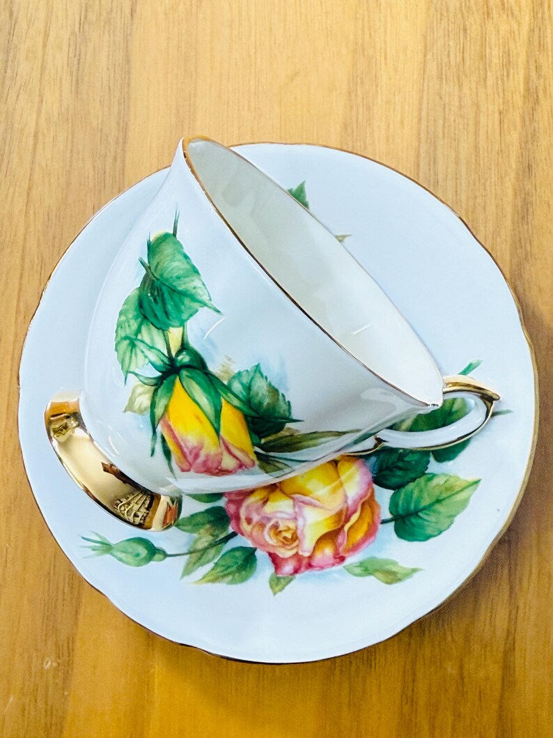 May include: White teacup and saucer set with a floral rose design in yellow and pink, accented with green leaves. The teacup and saucer have gold trim. The set is placed on a wooden surface.