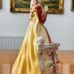 May include: A porcelain figurine of a woman in a long yellow dress with red sleeves, holding a small flower. The figurine is standing on a white balcony with floral accents. The woman has blonde hair and is looking to the side.
