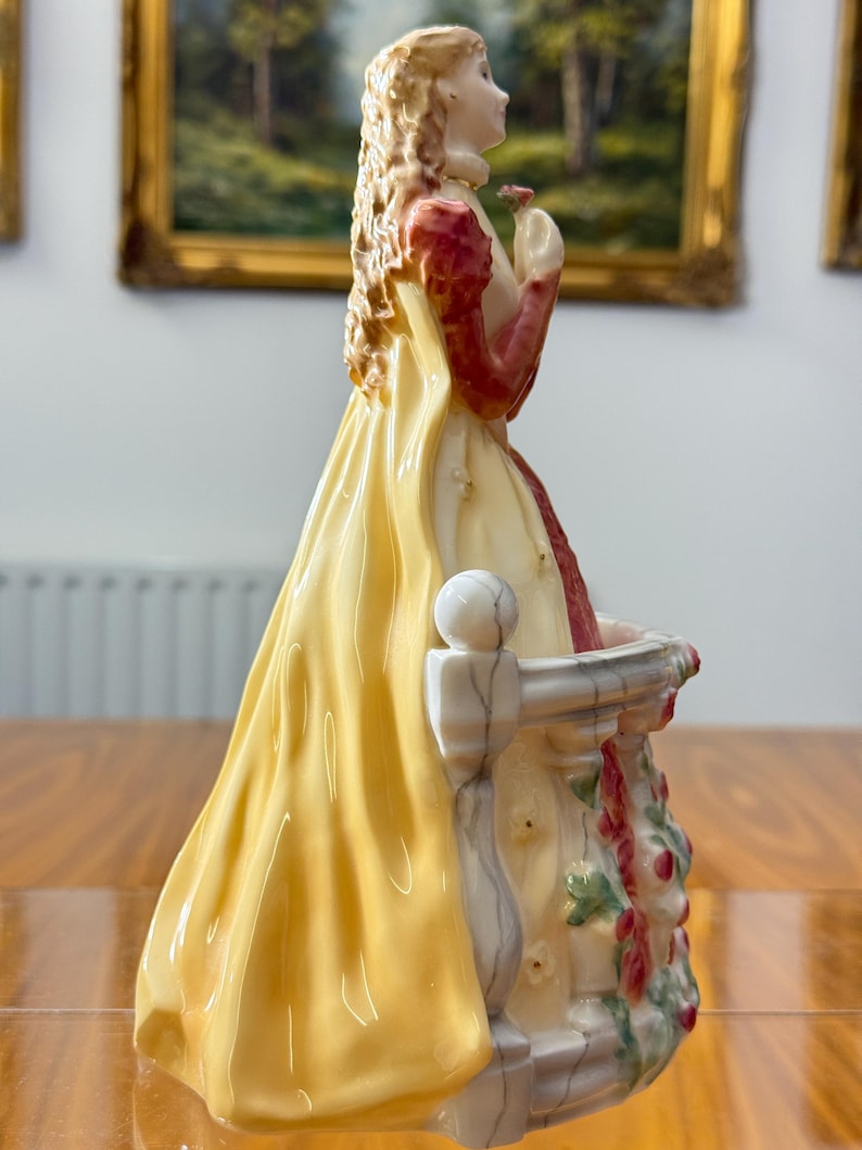 May include: A porcelain figurine of a woman in a long yellow dress with red sleeves, holding a small flower. The figurine is standing on a white balcony with floral accents. The woman has blonde hair and is looking to the side.