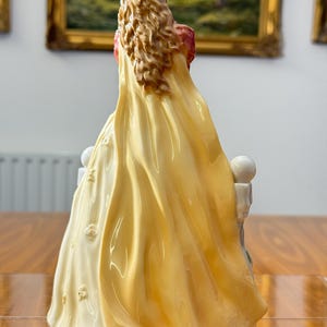 May include: A porcelain figurine of a woman in a long, flowing yellow gown with a red bodice. The figurine has long, wavy brown hair and is seated on a white structure with floral accents. The back view highlights the intricate details of the dress.