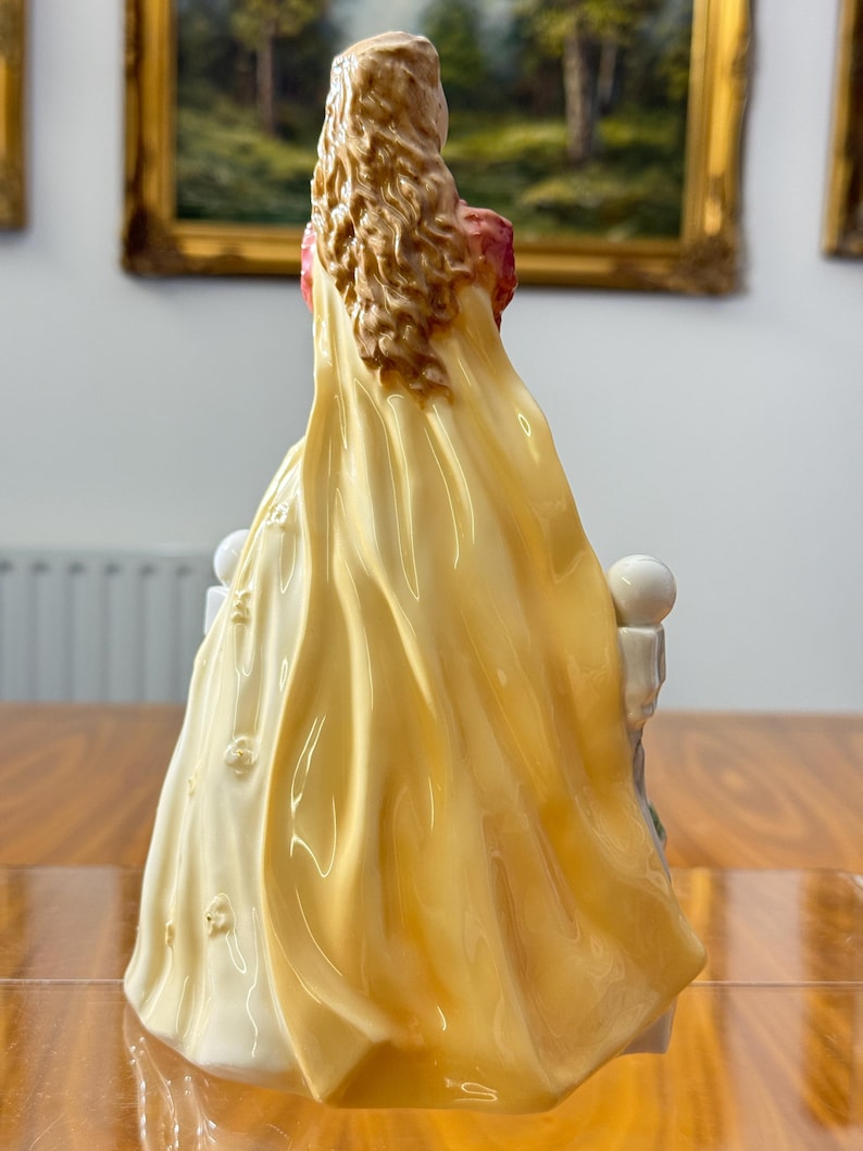 May include: A porcelain figurine of a woman in a long, flowing yellow gown with a red bodice. The figurine has long, wavy brown hair and is seated on a white structure with floral accents. The back view highlights the intricate details of the dress.