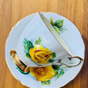 May include: A white teacup and saucer set with a yellow rose design. The teacup has a gold handle and trim, and the saucer has a matching floral pattern. The set is placed on a wooden surface.