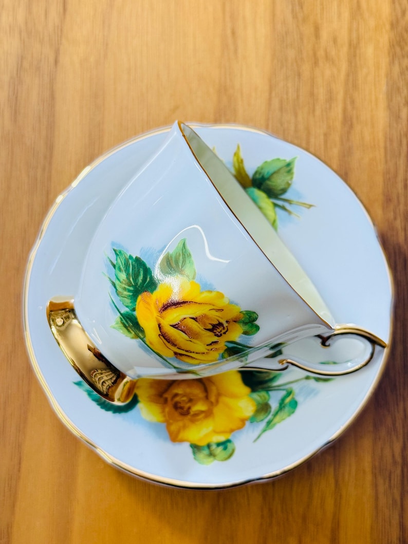 May include: A white teacup and saucer set with a yellow rose design. The teacup has a gold handle and trim, and the saucer has a matching floral pattern. The set is placed on a wooden surface.