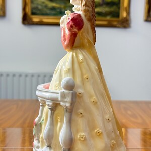 May include: A porcelain figurine of a woman in a long yellow dress with floral accents, holding a bouquet. The figurine has a white balcony with a floral design. The woman's sleeves are red, and her hair is blonde.