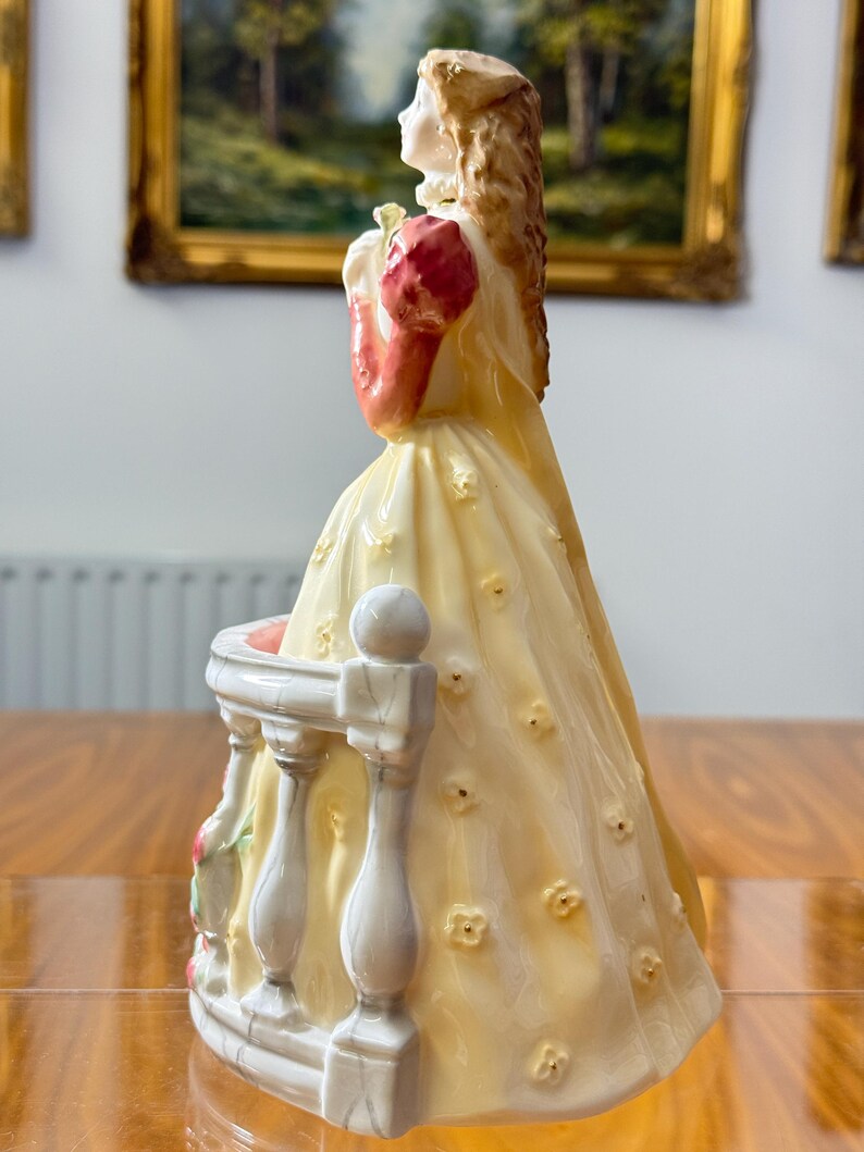 May include: A porcelain figurine of a woman in a long yellow dress with floral accents, holding a bouquet. The figurine has a white balcony with a floral design. The woman's sleeves are red, and her hair is blonde.