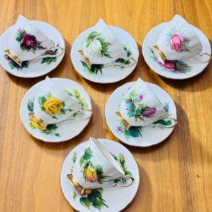 May include: Six white teacups and saucers with floral designs. Each cup and saucer features a different rose, in shades of red, pink, yellow, and white, with green leaves. The cups have gold-colored handles and rims. A classic tea set.