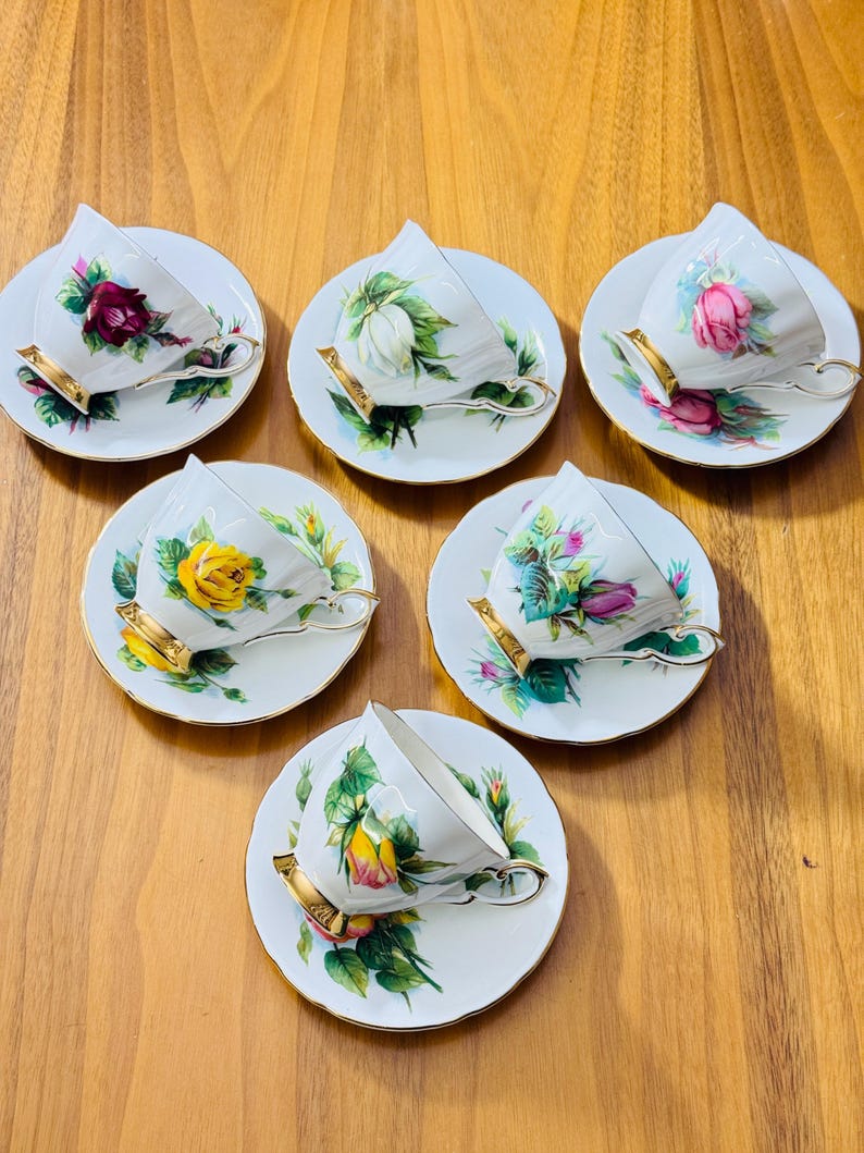 May include: Six white teacups and saucers with floral designs. Each cup and saucer features a different rose, in shades of red, pink, yellow, and white, with green leaves. The cups have gold-colored handles and rims. A classic tea set.