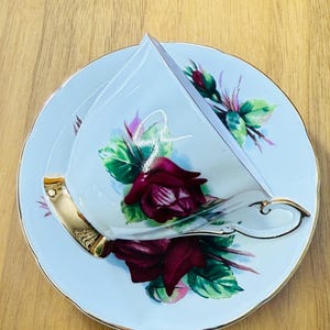 May include: A vintage teacup and saucer set. The light blue porcelain cup and saucer feature a burgundy rose design with green leaves and gold trim. The cup has a gold handle.