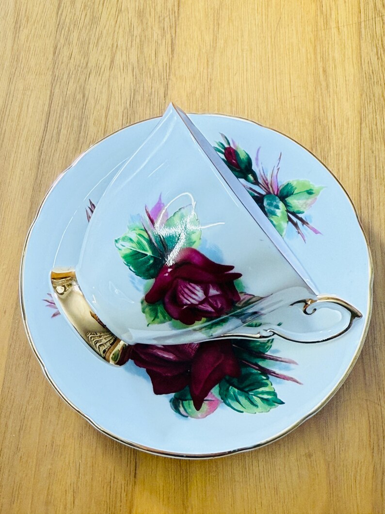 May include: A vintage teacup and saucer set. The light blue porcelain cup and saucer feature a burgundy rose design with green leaves and gold trim. The cup has a gold handle.