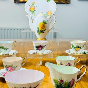 May include: A vintage tea set featuring a white teapot, serving plate, cups, creamer, and sugar bowl. The set is decorated with yellow and pink roses and gold trim. The tea set is displayed on a wooden table.