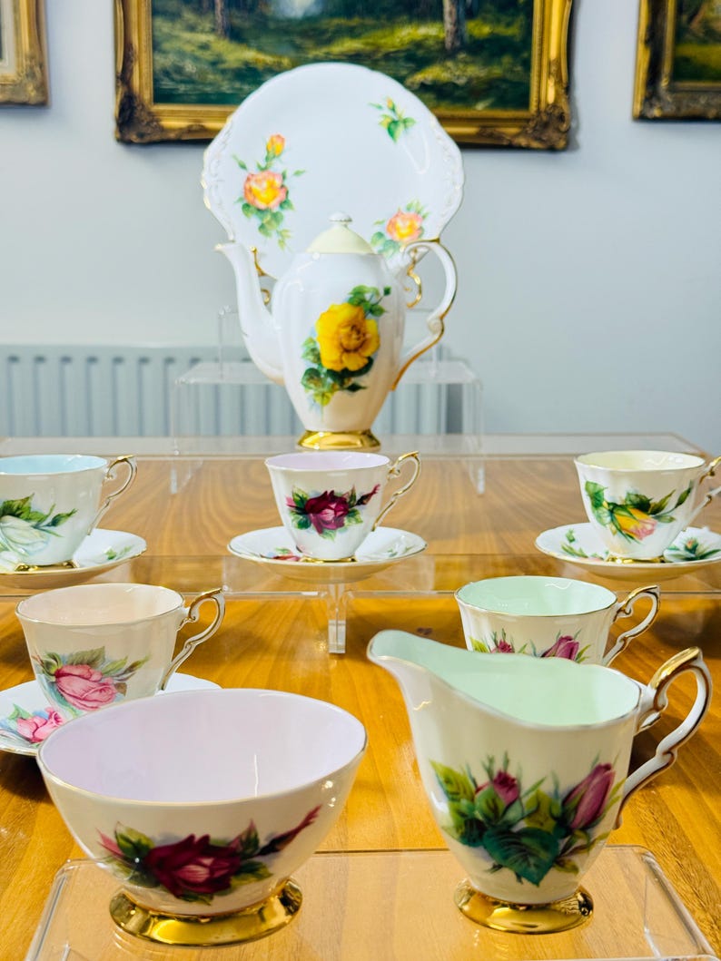 May include: A vintage tea set featuring a white teapot, serving plate, cups, creamer, and sugar bowl. The set is decorated with yellow and pink roses and gold trim. The tea set is displayed on a wooden table.