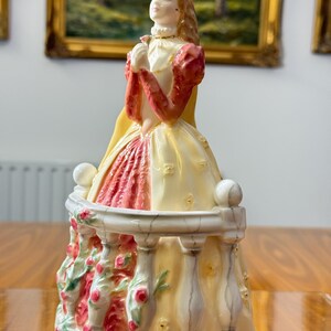 May include: A porcelain figurine of a woman in a long dress, standing on a balcony. The dress is cream and red, with a yellow cape. The balcony is decorated with roses. The woman has blonde hair and is holding a flower.