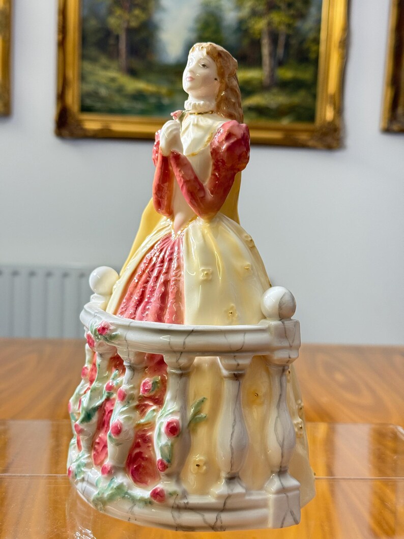 May include: A porcelain figurine of a woman in a long dress, standing on a balcony. The dress is cream and red, with a yellow cape. The balcony is decorated with roses. The woman has blonde hair and is holding a flower.