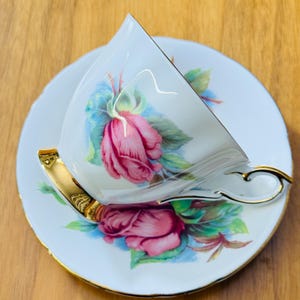 May include: White teacup and saucer set with a pink rose design and gold trim. The teacup is tilted, revealing the interior. The saucer and cup feature a floral pattern with green leaves and blue accents. The handle and rim of the cup are gold.