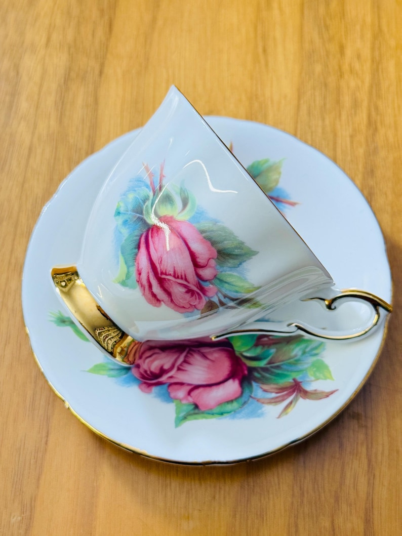 May include: White teacup and saucer set with a pink rose design and gold trim. The teacup is tilted, revealing the interior. The saucer and cup feature a floral pattern with green leaves and blue accents. The handle and rim of the cup are gold.