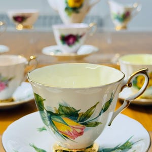 May include: A collection of vintage teacups and saucers with a floral rose design. The teacups have gold trim and handles, and the saucers have a matching floral pattern. A teapot and creamer are also visible in the background.
