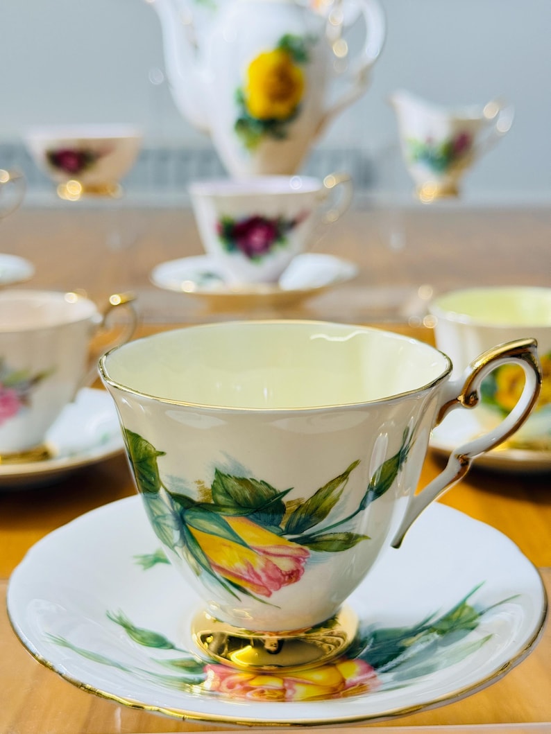 May include: A collection of vintage teacups and saucers with a floral rose design. The teacups have gold trim and handles, and the saucers have a matching floral pattern. A teapot and creamer are also visible in the background.