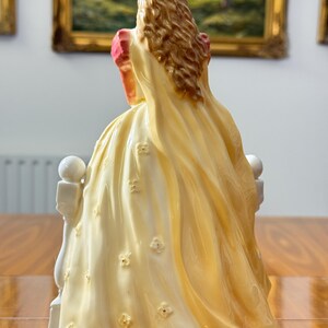 May include: A porcelain figurine of a woman in a long, flowing yellow dress adorned with small, raised floral details. The figurine has long, wavy brown hair and is seated on a white balustrade. The dress has a pink accent on the shoulder.