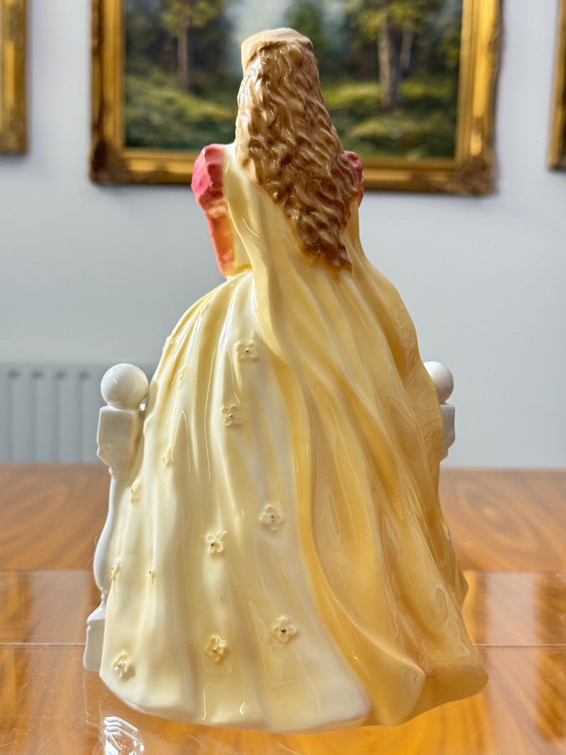 May include: A porcelain figurine of a woman in a long, flowing yellow dress adorned with small, raised floral details. The figurine has long, wavy brown hair and is seated on a white balustrade. The dress has a pink accent on the shoulder.