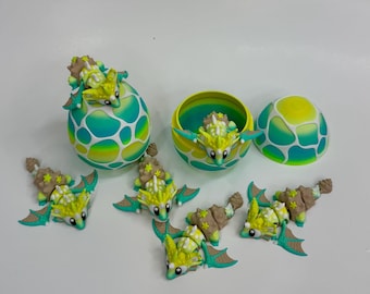 May include: A collection of fantasy-themed toys, including several small dragon figures with yellow, green, and brown coloring. The dragons have outstretched wings and star-shaped accents. Also included are two egg-shaped containers, one open and one closed, with a matching color scheme.