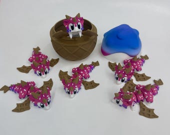 May include: A collection of whimsical dragon figurines in shades of pink, purple, and brown. The dragons have waffle-like wings and are scattered around a brown egg-shaped container and a blue and white blob.