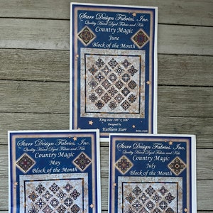 Op de afbeelding: Drie bedrukte stoffen kits voor een quilt, elk met een gedetailleerd patchwork ontwerp in blauw, bruin en crème tinten. De kits zijn gelabeld "Country Magic" en "Block of the Month" voor mei, juni en juli. Elke kit is king size, 269 cm x 269 cm.
