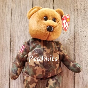 Pode incluir: Um urso beanie de pelúcia marrom com camuflagem com um pequeno patch da bandeira americana no braço e a palavra "Peaknits" impressa no peito. O urso está vestindo um traje de camuflagem e tem uma etiqueta Ty presa na orelha.