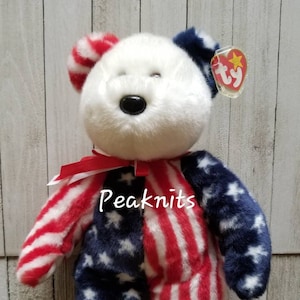 May include: A plush teddy bear with patriotic colors. The bear features a white head, red and white striped arms and legs, and blue sections with white stars. A red ribbon is tied around the neck, and a Ty tag is attached.