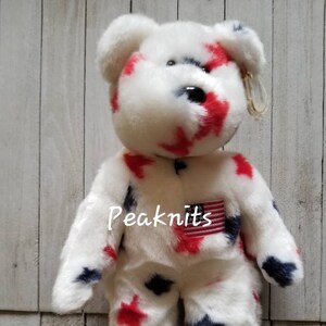 Puede incluir: Oso de peluche blanco con estrellas rojas y azules y un pequeño parche de bandera estadounidense. El oso es un Beanie Baby.