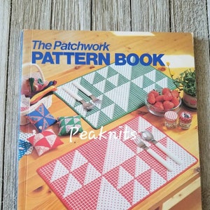 以下が含まれることがあります： 「The Patchwork PATTERN BOOK」というタイトルの本。赤い白の組み合わせのパッチワークのランチョンマットと緑と白の組み合わせのパッチワークのランチョンマットが木製のテーブルの上に置かれている。ランチョンマットは白い水玉模様で飾られている