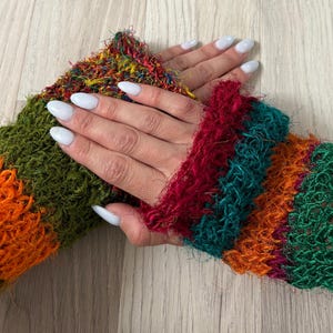 May include: A pair of fingerless gloves made from a fuzzy, textured yarn featuring horizontal stripes in vibrant shades of orange, olive green, deep red, and teal. The knit construction has a loose, open weave that creates a soft, shaggy appearance.