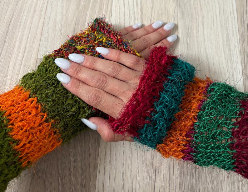 May include: A pair of fingerless gloves made from a fuzzy, textured yarn featuring horizontal stripes in vibrant shades of orange, olive green, deep red, and teal. The knit construction has a loose, open weave that creates a soft, shaggy appearance.