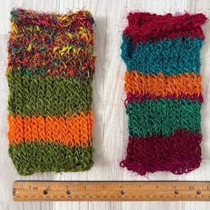 May include: Two rectangular scrubbers with a textured, loopy knit construction. The left scrubber features a multicolored top section with green and orange stripes below. The right scrubber has horizontal stripes of maroon, teal, and orange. A 10-inch ruler is visible.