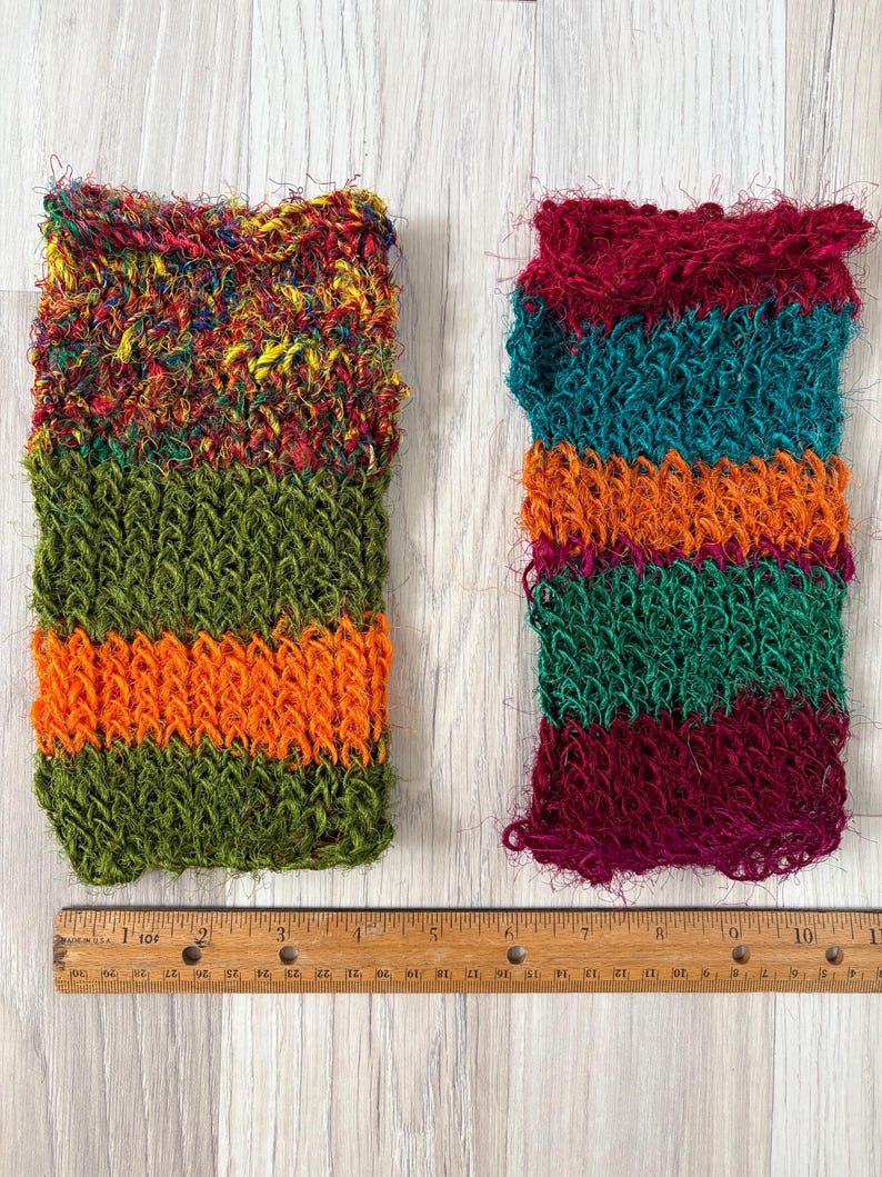 May include: Two rectangular scrubbers with a textured, loopy knit construction. The left scrubber features a multicolored top section with green and orange stripes below. The right scrubber has horizontal stripes of maroon, teal, and orange. A 10-inch ruler is visible.