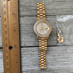 May include: A gold-tone watch with a white face and diamond accents. The watch has a gold-tone band with diamond accents. The watch is on a wooden surface.