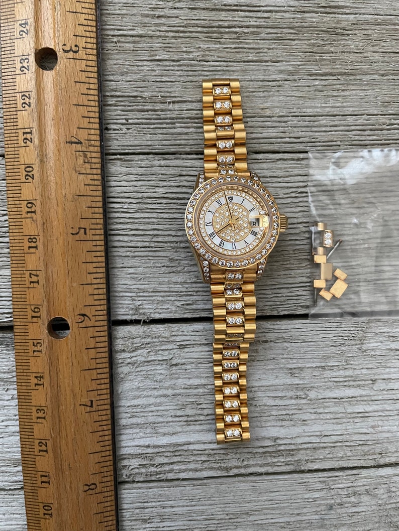May include: A gold-tone watch with a white face and diamond accents. The watch has a gold-tone band with diamond accents. The watch is on a wooden surface.