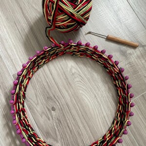 May include: A circular knitting loom with a woven yarn design in red, black, yellow, and green. A ball of matching yarn and a crochet hook with a wooden handle are also present, suggesting a crafting project.