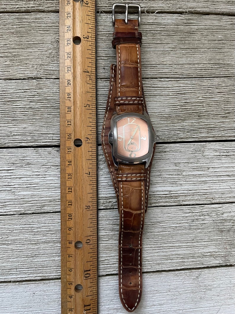 May include: A brown leather wristwatch with a silver case and a white face. The watch has a white stitching detail on the band.