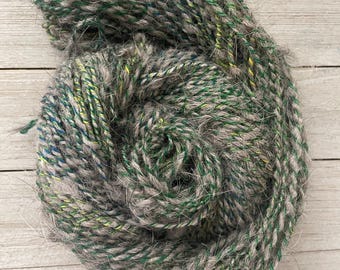 May include: A skein of yarn in shades of gray, green, and blue. The yarn is made of a blend of wool and mohair, giving it a soft and fuzzy texture.