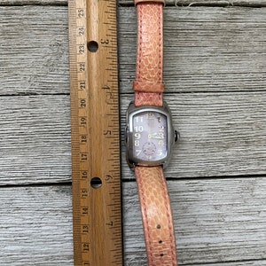 May include: A silver wristwatch with a pink leather band. The watch face is white with black Roman numeral hour markers and black hands. The watch is about 2 inches in diameter.